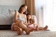 © sementsova321 - Positive young adult Caucasian female wearing casual style clothing posing indoor in living room with her daughter while sitting on the floor near sofa and window, watching funny video with child.