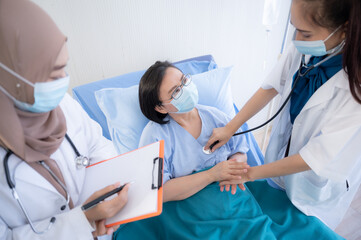 Wall Mural - Portrait of two beautiful and cheerful female doctors wearing labcoat and stethoscope embracing senior patient sitting on hospital bed looking at camera