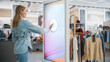 Beautiful Female Customer Using Floor-Standing LCD Touch Display while Shopping in Clothing Store. She is Pressing Button to Start Using a Screen. People in Fashionable Shop.
