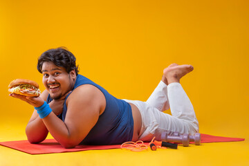  A fat man lying on yoga mat holding a burger with dumbles and skipping rope kept beside.