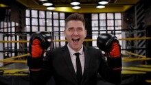 Portrait Of A Successful Businessman In Boxing Gloves And A Suit In A Boxing Ring, He Jumps With Happiness And Laughs Looking At The Camera.