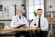 © sofiko14 - Portrait of caucasian man in business clothes sitting at desk with aged doctor that writing on clipboard. Therapist and patient looking at camera. Health examination concept.