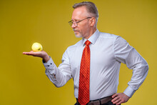 Mature Man Looking At An Apple And Posing To The Camera. Male In White Shirt And Red Tie. Yellow Background