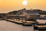 Fototapeta Na ścianę - A ferry is moored at the terminal in the Spanish port of Ibiza. It is a summer morning and behind the ferry the morning sun is in the orange sky. A flock of birds flies by.