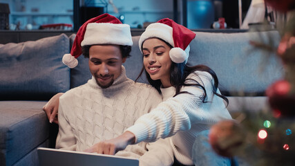 Wall Mural - happy african american couple in santa hats using laptop near blurred christmas tree