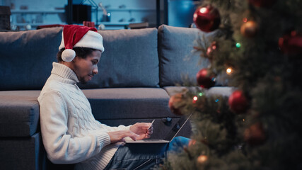 Wall Mural - side view of african american freelancer in santa hat using laptop near blurred christmas tree