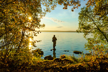 A Fisherman Stands On A Rock Near The Shore Of The Lake.