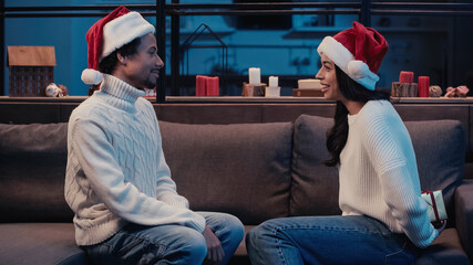 Wall Mural - african american woman hiding christmas present near happy boyfriend in santa hat