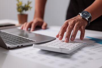 Business background in tax, accounting, statistics, and analytic research concept, a businessman uses a calculator and laptop to do math finance on a white desk in his office.