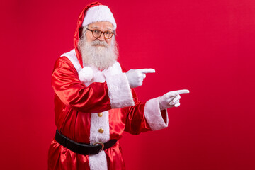 Funny happy excited old bearded Santa Claus face wearing costume looking at camera showing pointing fingers aside advertising Christmas promotion, New Year xmas discount ad isolated on red background.