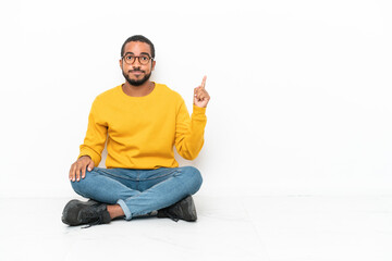 Wall Mural - Young Ecuadorian man sitting on the floor isolated on white wall pointing with the index finger a great idea