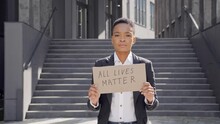 Black Girl With A Stylish Hairstyle Holding Carton Placard With All Lives Matter Writing. Female Activist Protesting Against Racism While Standing At Street Near Politic Buidings. Concept Of Equality.