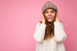 © Ivan Traimak - Photo of beautiful happy smiling young brunette woman isolated over pink background wall wearing knitted white sweater and grey warm hat looking at camera
