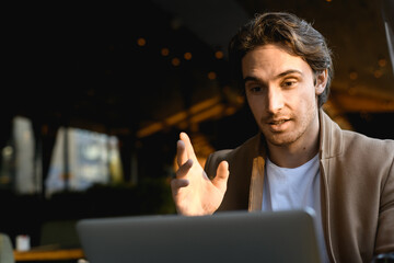 Good looking freelancer in coat having video call on laptop on terrace of cafe 