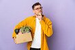 Young caucasian man making a move while picking up a box full of things isolated on purple background having doubts and thinking
