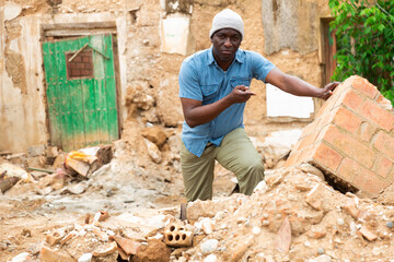 Portrait of upset adult african man standing in front of old ruined house after hurricane outdoors