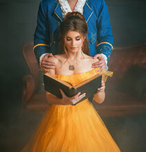 Woman Princess Holding A Book Without Title Cover Design Reads The Text. Fantasy Man Enchanted Prince Hugging A Beautiful Lady By Shoulders. Girl In Yellow Medieval Historical Dress Vintage Gown.