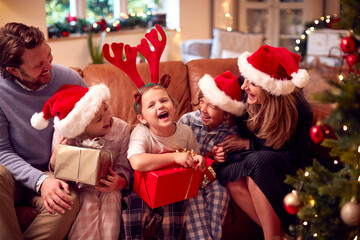 Wall Mural - Family Wearing Santa Hats Sitting On Sofa At Home Opening Christmas Gifts