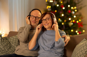 Wall Mural - winter holidays, technology and people concept - happy senior couple with headphones listening to music at home in evening over christmas tree lights on background
