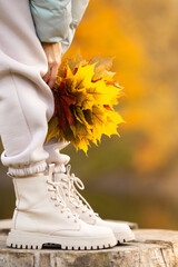 Close up of female legs wearing trendy beige boots with bouquet of colorful maple leaves. Autumn mood. Stylish shoes, autumn fashion, autumn sale. Vertical image. Selective focus.