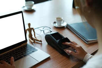 Behind shot of a young woman is using a credit card reader machine and white blank screen computer laptop at the wooden cashier.