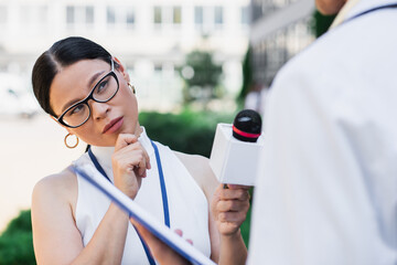 Poster - brunette asian journalist holding microphone and looking at blurred doctor in white coat