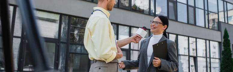 Poster - newscaster holding microphone and taking interview of cheerful asian businesswoman in glasses standing near building, banner