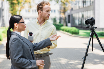 Canvas Print - reporter holding microphone near asian businesswoman pointing at digital camera on tripod