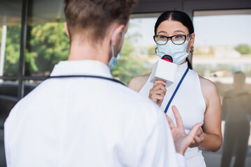 Wall Mural - asian reporter in glasses and medical mask talking in microphone near blurred doctor in white coat