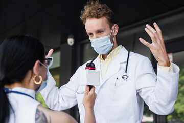 Poster - doctor in medical mask gesturing during interview with blurred asian reporter