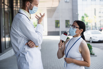 Wall Mural - side view of asian reporter in medical mask talking in microphone while taking interview of doctor near clinic