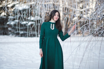 Friendship of an owl and a girl. Myth, fairy tale. Mystical spirit. Fantasy female elf walks with an owl in the winter forest. Beautiful brunette brunette girl in a green vintage dress. Tame bird.