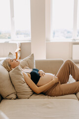 Wall Mural - Pregnant woman wearing cozy home wear, lying on a sofa in a light interior, holding hands on belly.