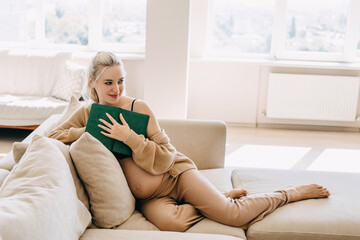 Wall Mural - Blonde pregnant woman wearing home wear, sitting on a sofa at home, holding a book.