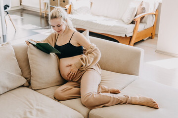 Wall Mural - Blonde pregnant woman wearing home wear, sitting on a sofa at home, reading a book.