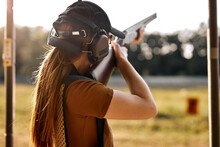 Young Caucasian Woman On Tactical Gun Training Classes. Woman With Weapon, Wearing Cap, Protective Headphones And Eyeglasses. Outdoor Shooting Range. At Sunny Evening At Summer. Rear View