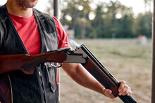 Cropped Man In Uniform Charges Pump-action Shotgun In An Outdoor Range. Skilled Experienced Man Is Preparing To Shoot. Firearms For Sports Shooting, Hobby. Copy Space. Close-up Hands, Smoke From Rifle