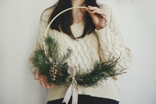 Stylish Woman In Cozy Sweater Holding Modern Christmas Wreath On White Background Indoors. Atmospheric Image. Winter Holiday Preparation And Decor. Making Boho Christmas Wreath