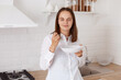 © sementsova321 - Portrait of smiling attractive young adult woman with dark hair wearing white casual shirt, having breakfast in the kitchen, holding plate in hands, looking at camera with pleasant expression.