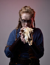 Closeup Portrait Of Young Red Haired Woman With Black Barbarian Facepaint Wearing Medieval Viking Inspired Costume Holding  Sheep Skull For Ritual  Posing Against Studio Background With Red Lighting