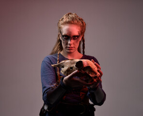 Closeup portrait of young red haired woman with black barbarian facepaint wearing medieval viking inspired costume Holding  sheep skull for ritual  posing against studio background with red lighting