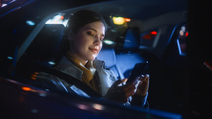 Wall Mural - Stylish Female is Commuting Home in a Backseat of a Taxi at Night. Beautiful Woman Passenger Using Smartphone while in a Car in Urban City Street with Working Neon Signs.