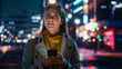 © Gorodenkoff - Portrait of a Beautiful Woman in Trench Coat Walking in a Modern City Street with Neon Lights at Night. Attractive Female Using Smartphone and Looking Around the Urban Cinematic Environment.