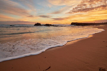 A beautiful beach sunset illuminating the sky with vibrant orange colours, a scenic view of the ocean with waves washing onto the shore along the coast of Phillip Island, Australia.
