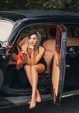Vintage Style Photo. Elegant Retro Woman In Black Evening Long Dress Posing Sits In Old Car On Road, Summer Nature Background Green Leaves Trees. Red Lips Shoes Headscarf. Beautiful Face Bright Makeup