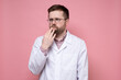 © koldunova - Caucasian doctor in a white coat pensively and anxiously looks to the side, holding hand to face. Pink background.