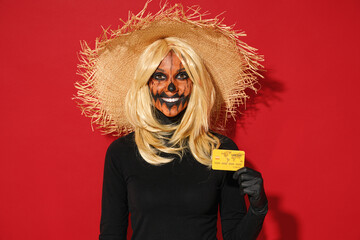 Young fun woman with Halloween makeup mask wears straw hat black scarecrow costume hold in hand credit bank card isolated on plain red background studio portrait. Celebration holiday party concept