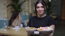 Portrait Of Young Positive Caucasian Waitress Posing With Seafood On Tray Looking At Camera Smiling. Slim Beautiful Woman Standing In Restaurant With Order And Blurred Customers At Background