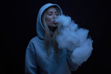 Wall Mural - Portrait of beautiful blonde girl in gray hoodie smokes vape isolated on black studio background, cloud of steam smoke, mini hookah