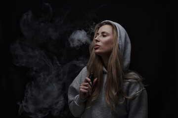 Wall Mural - Portrait of beautiful blonde girl in gray hoodie smokes vape isolated on black studio background, cloud of steam smoke, mini hookah